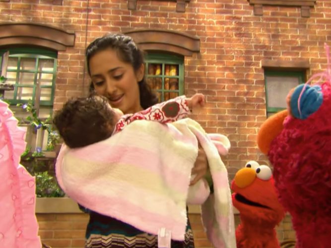 A grown-up holding their baby while Elmo looks on.