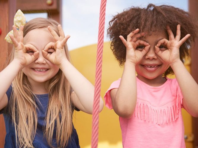 Two young children look through googles they've made by forming rings with their index fingers and thumbs.