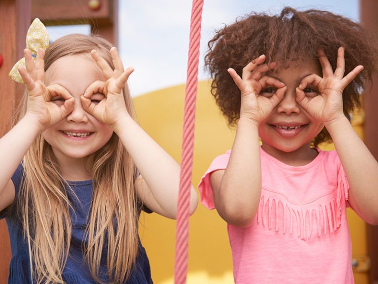 Two young children look through googles they've made by forming rings with their index fingers and thumbs.