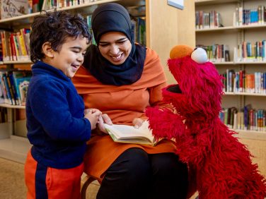 A woman in a children's library reading to Elmo and a child.