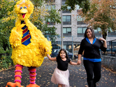 Big Bird walks towards us, holding the hand of a young child who's holding the hand of an adult.